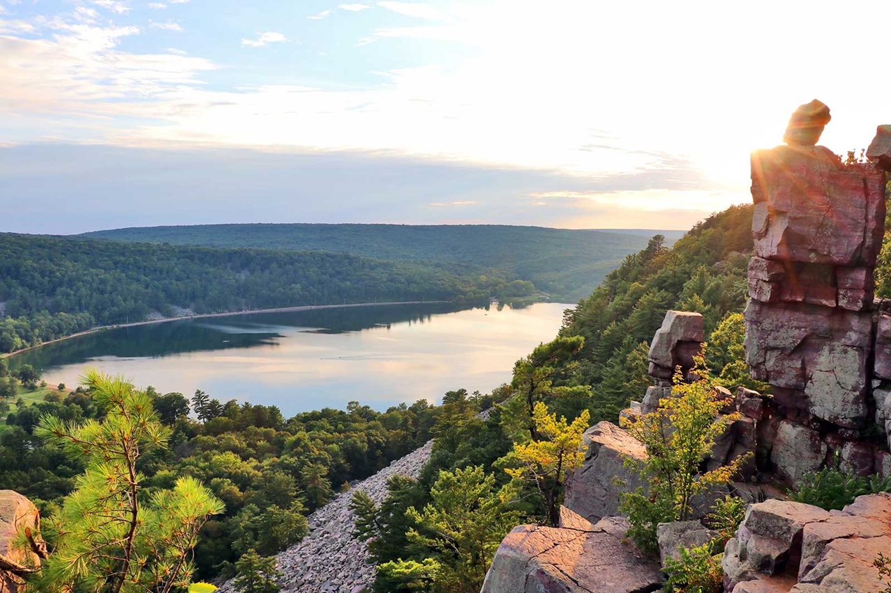 A scenic view of a lake surrounded by lush trees, with rocky cliffs illuminated by sunlight during sunset.