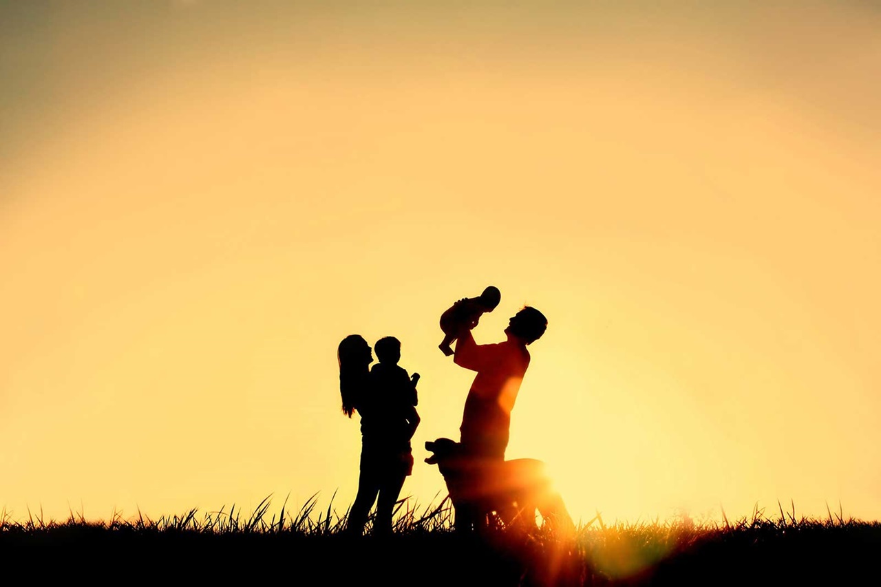 A silhouetted family at sunset, with parents holding children and a dog nearby in a grassy field.