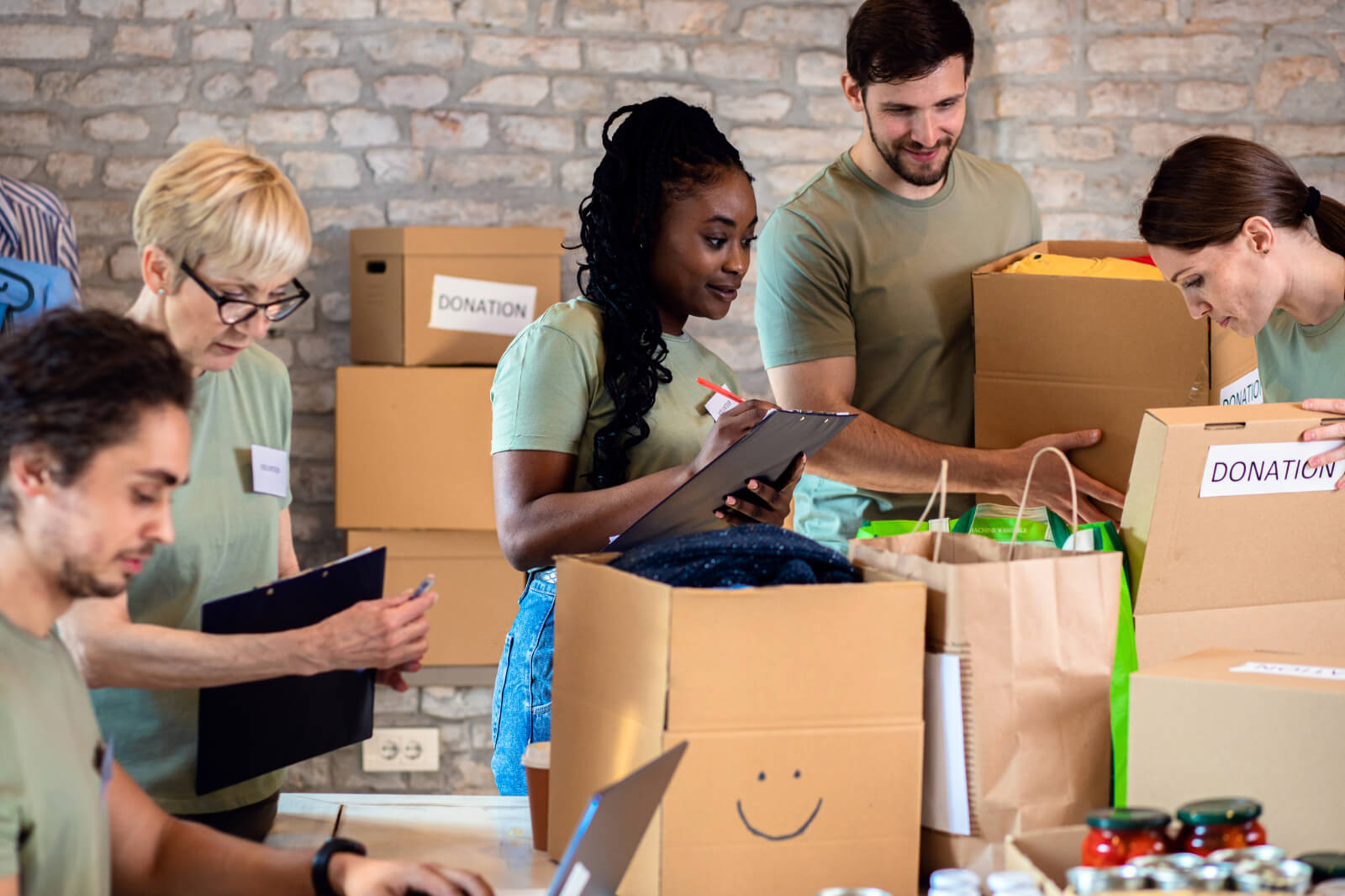A group of volunteers sorting donations in a bright room with stone walls. They handle boxes labelled 'DONATION' and clipboards.
