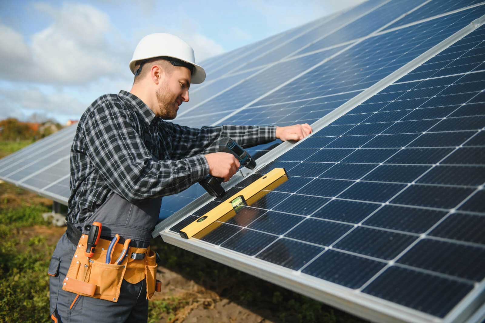 A worker in a hard hat installs solar panels, using a drill and a level on a sunny day, surrounded by solar arrays.