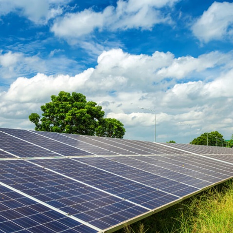 A solar panel array under a blue sky with scattered clouds and green trees nearby.