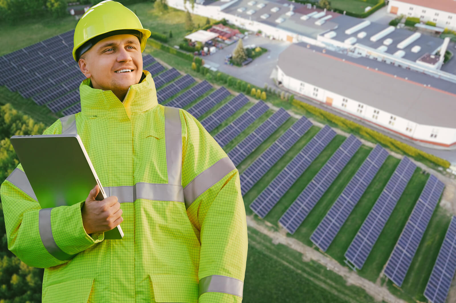 A person in a high-visibility jacket stands near a large solar panel field and a building in the background.