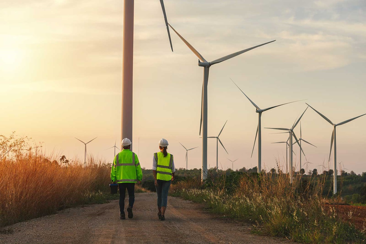 Two workers in high-visibility jackets walking along a dirt path surrounded by wind turbines at sunset.