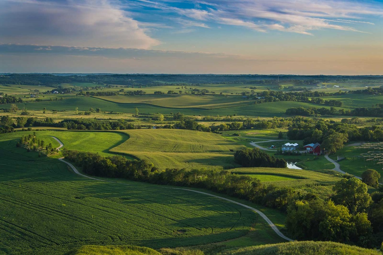 A panoramic view of lush green fields with a winding road, a red barn, and a serene pond under a clear blue sky.