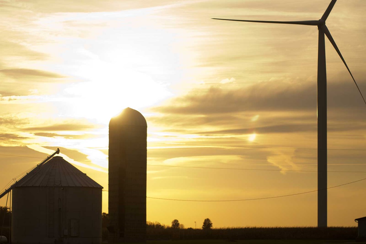 A sunset silhouette of a wind turbine and silos, with warm hues illuminating the sky and landscape.