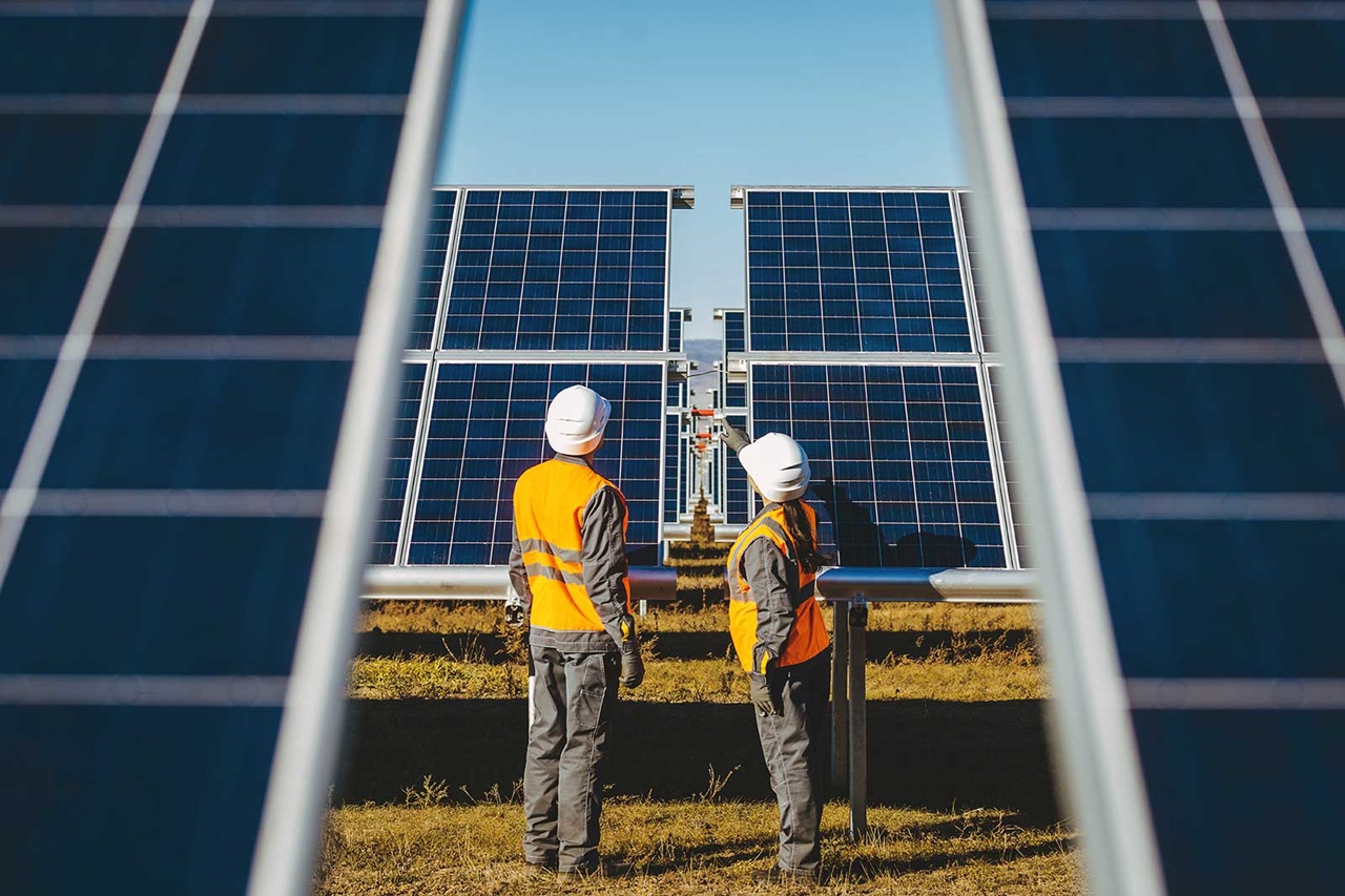 Two workers in safety gear examine solar panels in a field under a clear blue sky.