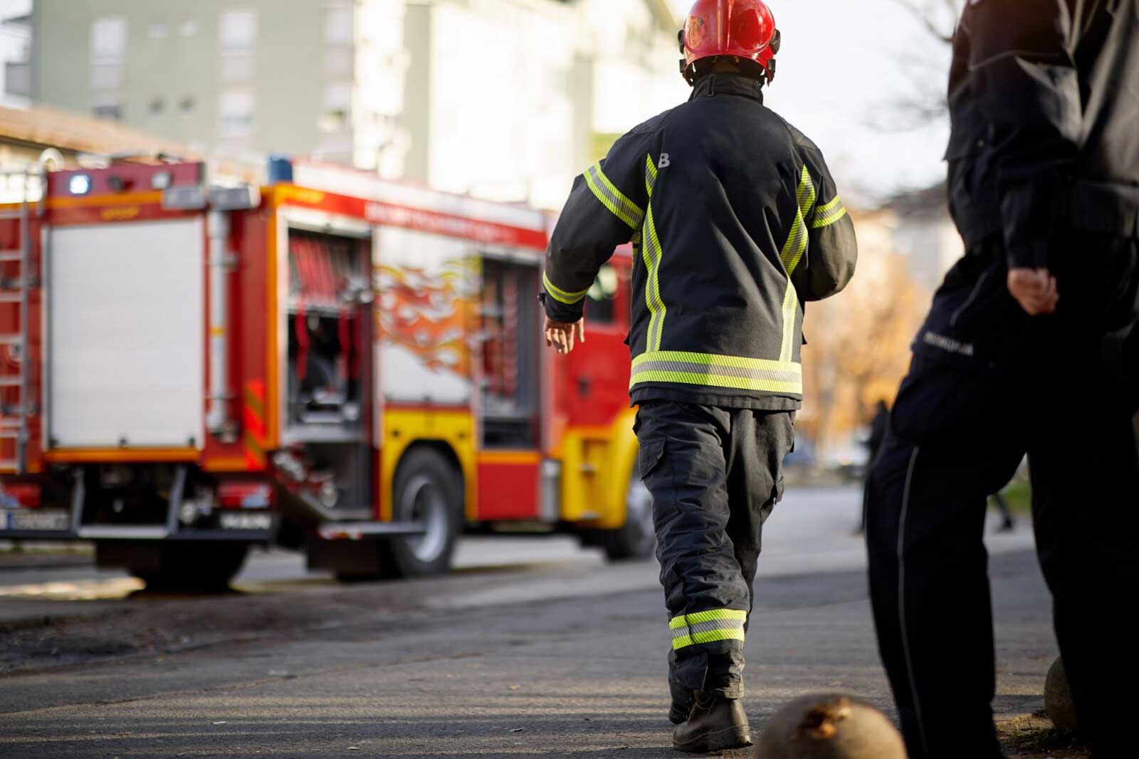 A firefighter in uniform walks towards a fire truck, with another person slightly visible to the right.