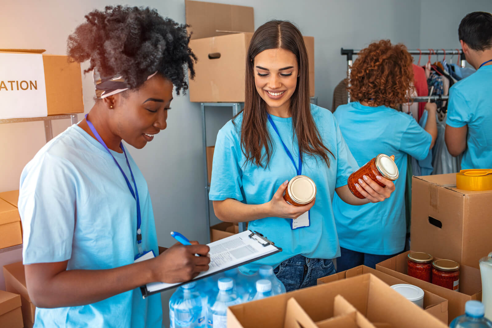 Volunteers in blue shirts work in a donation centre, packaging jars and taking notes among cardboard boxes.