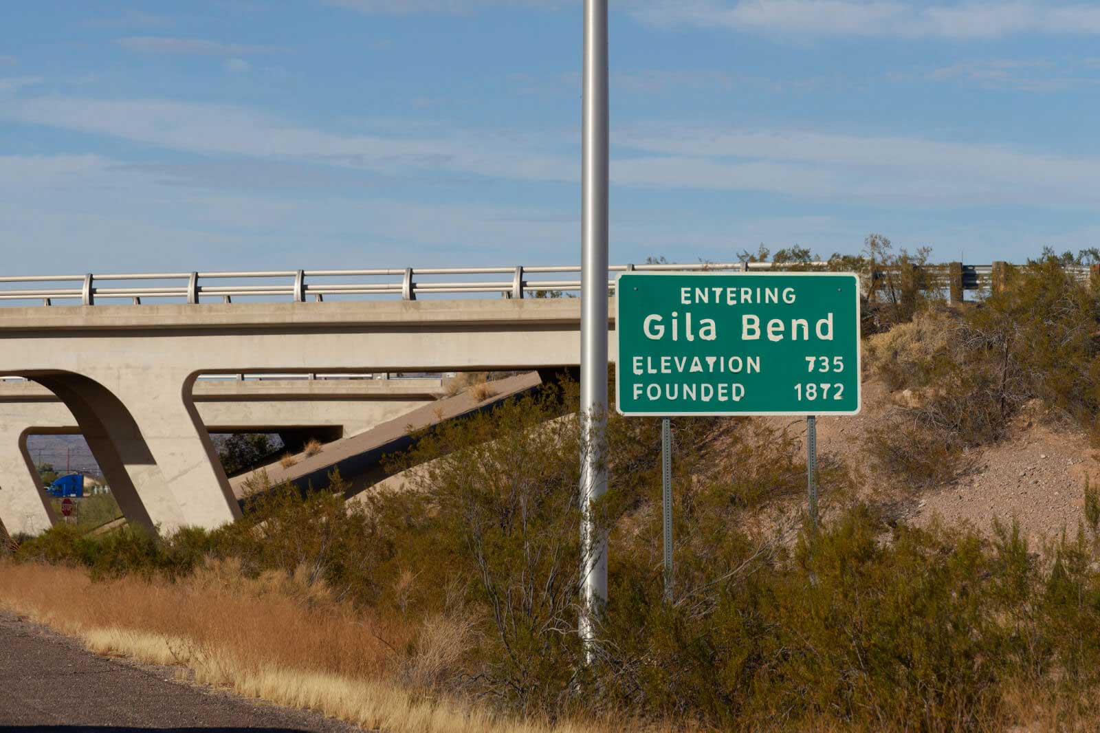 A road sign indicating entry into Gila Bend, elevation 735 feet, founded in 1872, near a highway bridge.