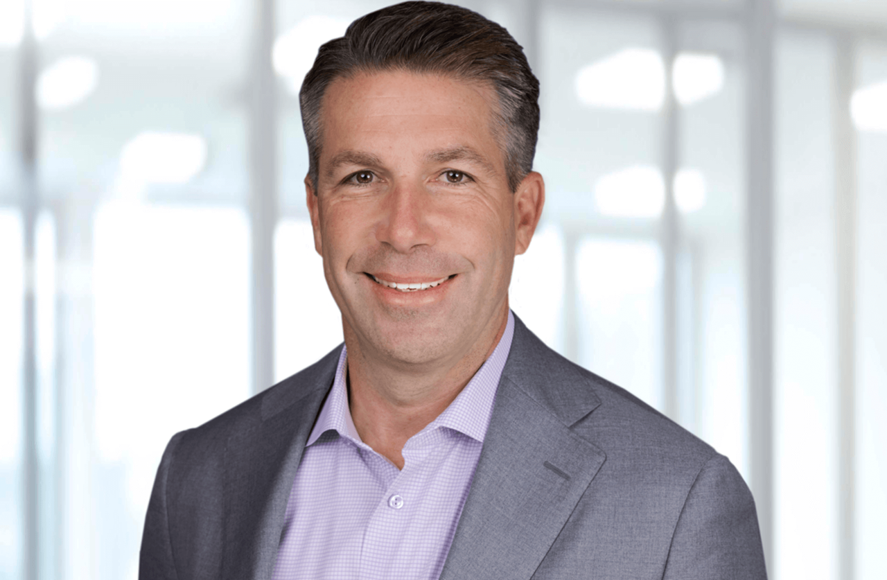 A professional man in a grey suit and checked shirt stands against a blurred office background.