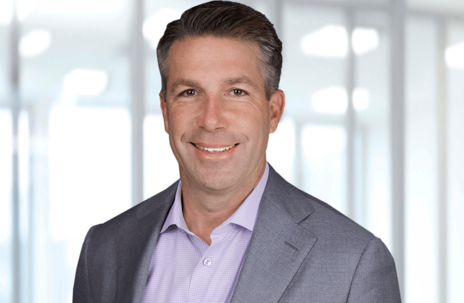 A professional man in a grey suit and checked shirt stands against a blurred office background.