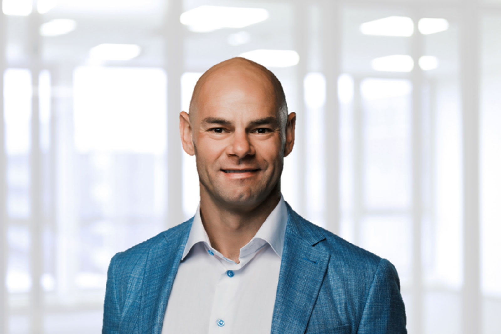 A man in a blue blazer stands in a bright office with large windows, highlighting a professional setting.