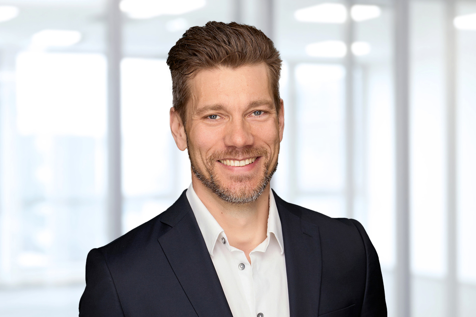A man in a black suit and white shirt stands against a softly blurred office background.