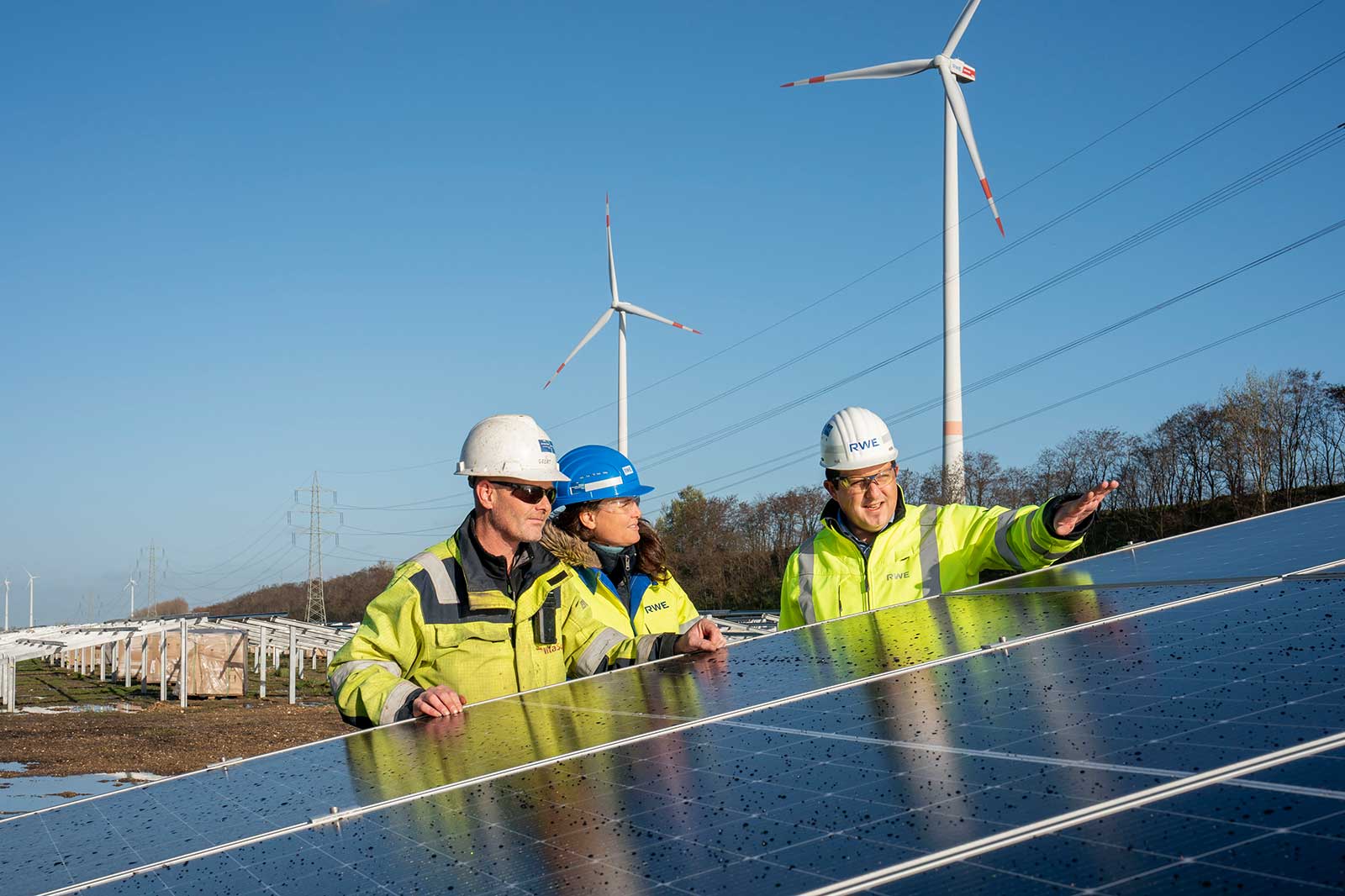 Professionals inspect a solar panel with wind turbines in the background under a blue sky.