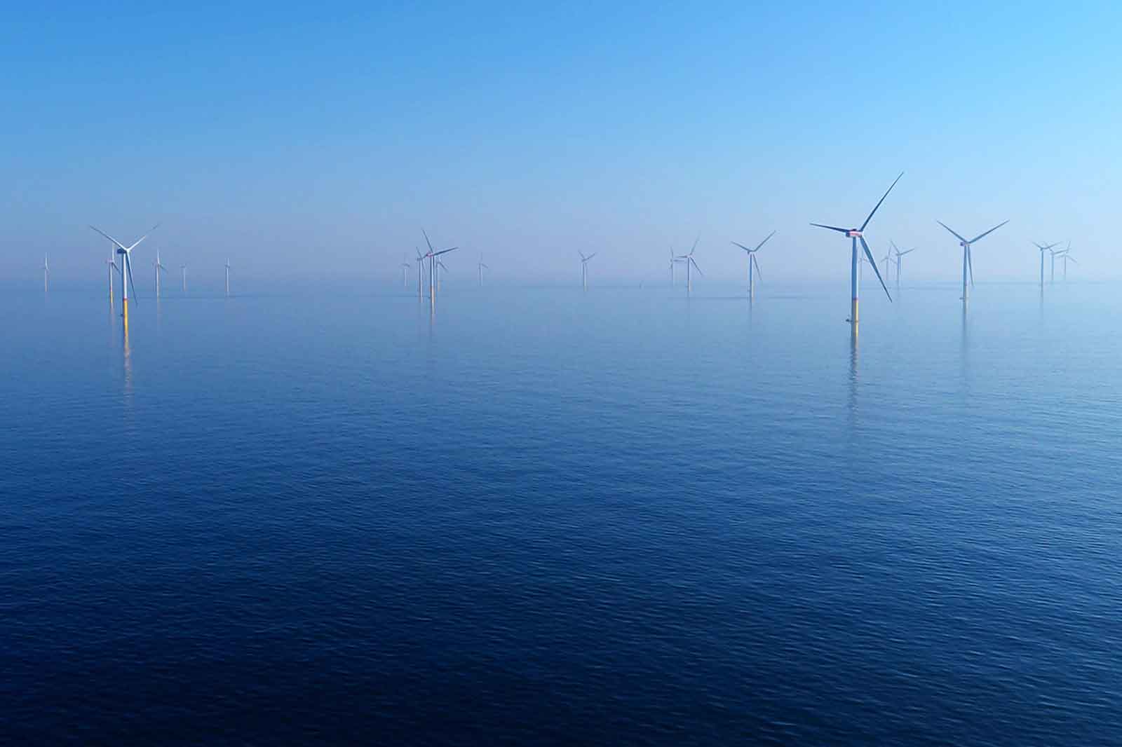 Wind turbines stand in the water against a calm blue surface and a clear blue sky in the background.