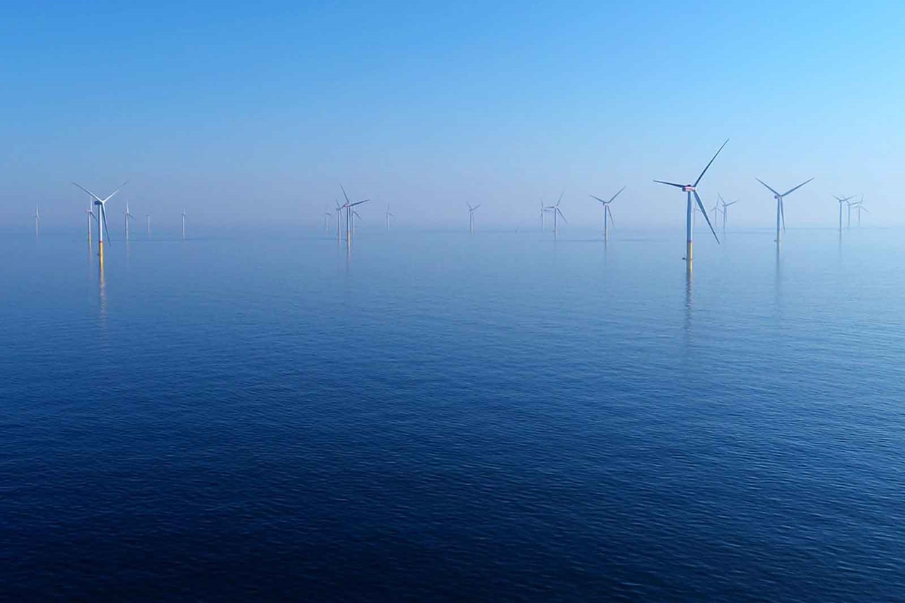 Wind turbines stand in the water against a calm blue surface and a clear blue sky in the background.