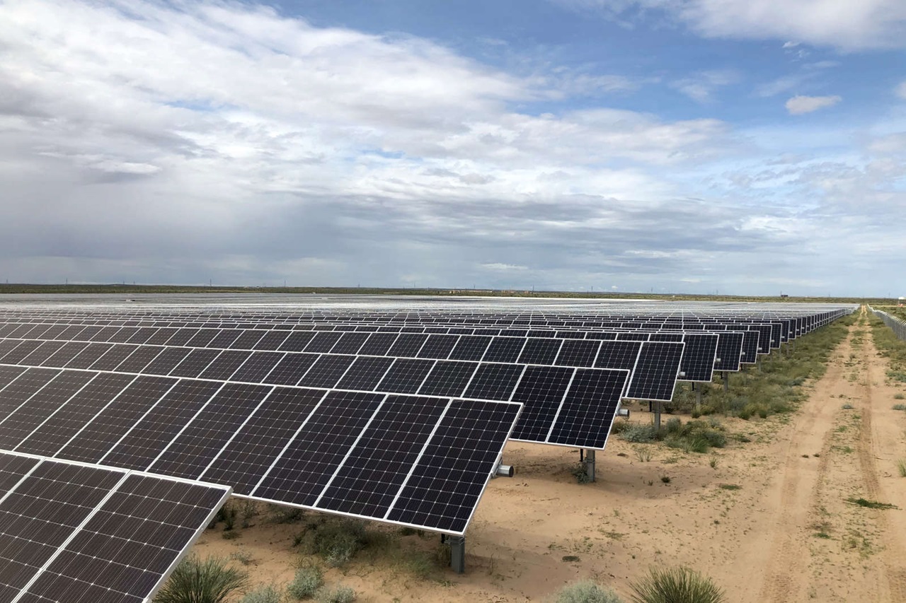 A wide-angle image of a solar farm featuring numerous solar panels in an open, sandy landscape beneath a cloudy sky.