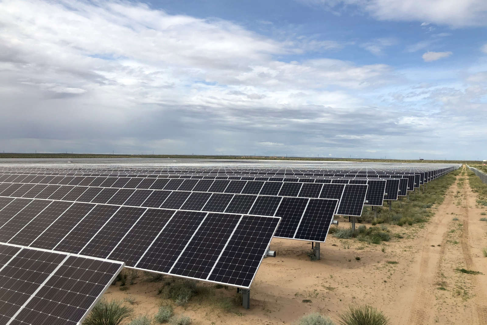 A wide-angle image of a solar farm featuring numerous solar panels in an open, sandy landscape beneath a cloudy sky.