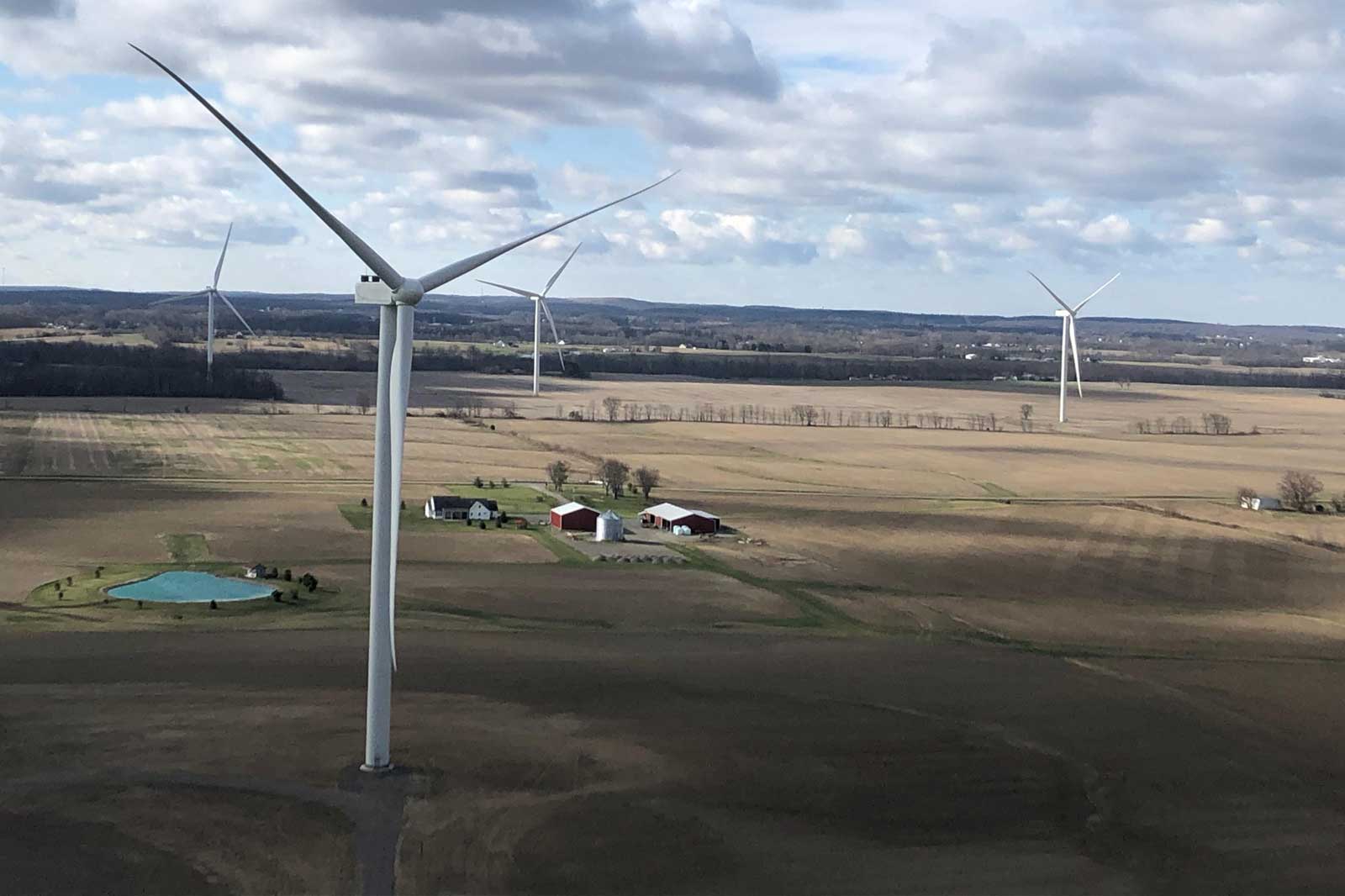 A vast field featuring wind turbines and a farmhouse. Clouds fill the sky.