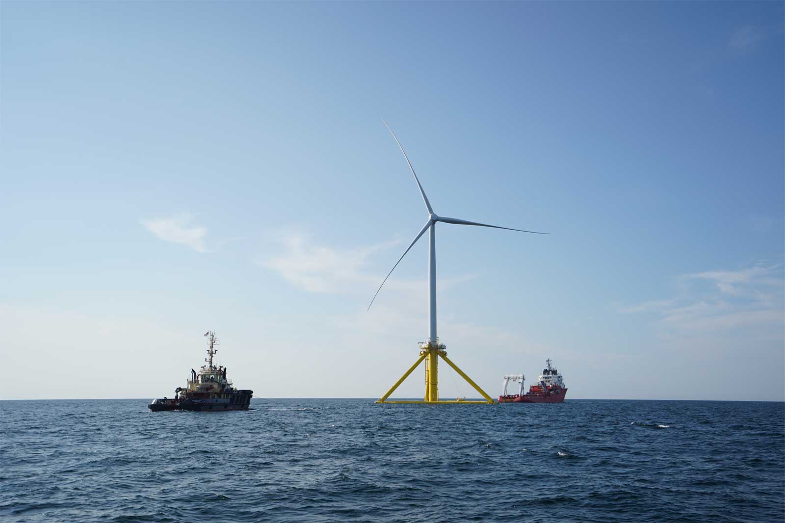 A wind turbine stands in the water, surrounded by two service boats under a clear sky.