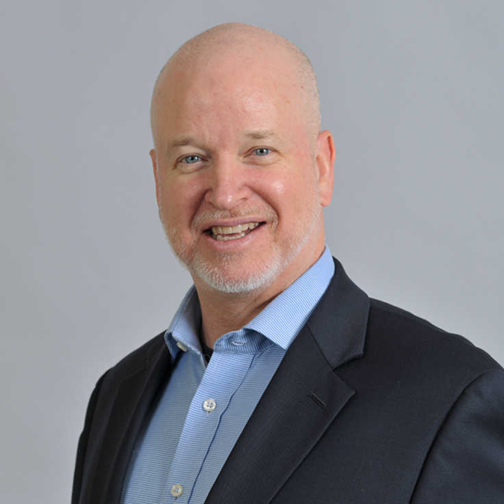 A man in a business suit with a light blue shirt, set against a grey background.