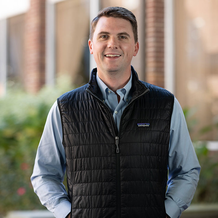 A man in a black vest stands outdoors, wearing a light blue shirt, with greenery in the background and soft natural lighting. Portrait of Ryan Ferguson.