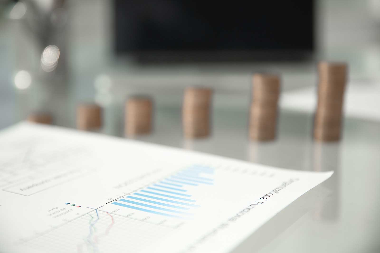 A close-up of a financial report on a glass table, with blue bar graphs and stacks of coins in the background.
