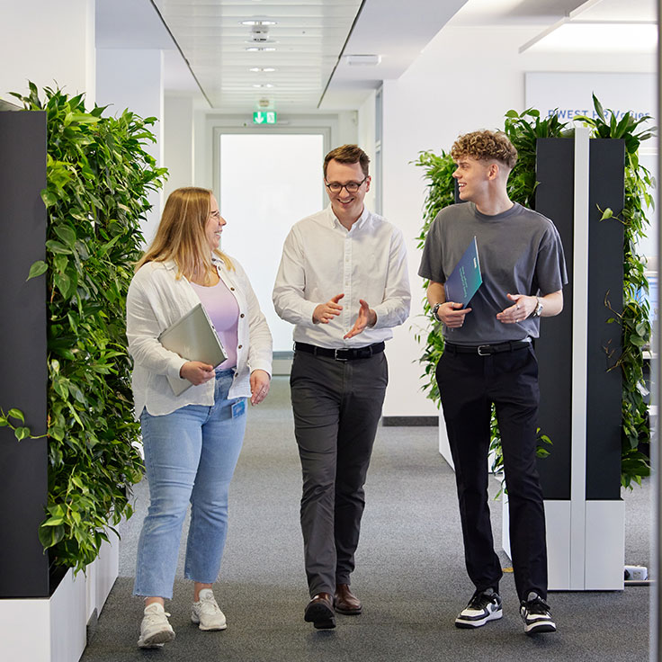 Three colleagues are walking and talking in a modern office corridor, surrounded by greenery.