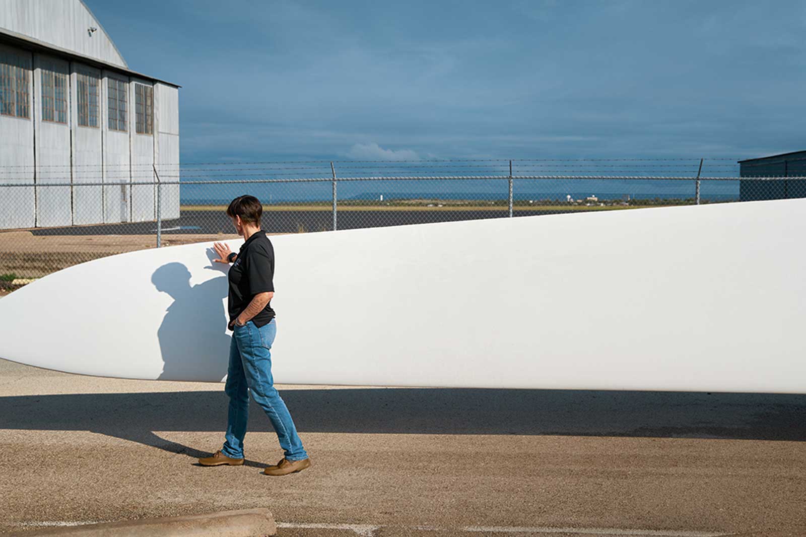 A person walks beside a large wind turbine blade, touching its surface, with a hangar and a blue sky in the background.