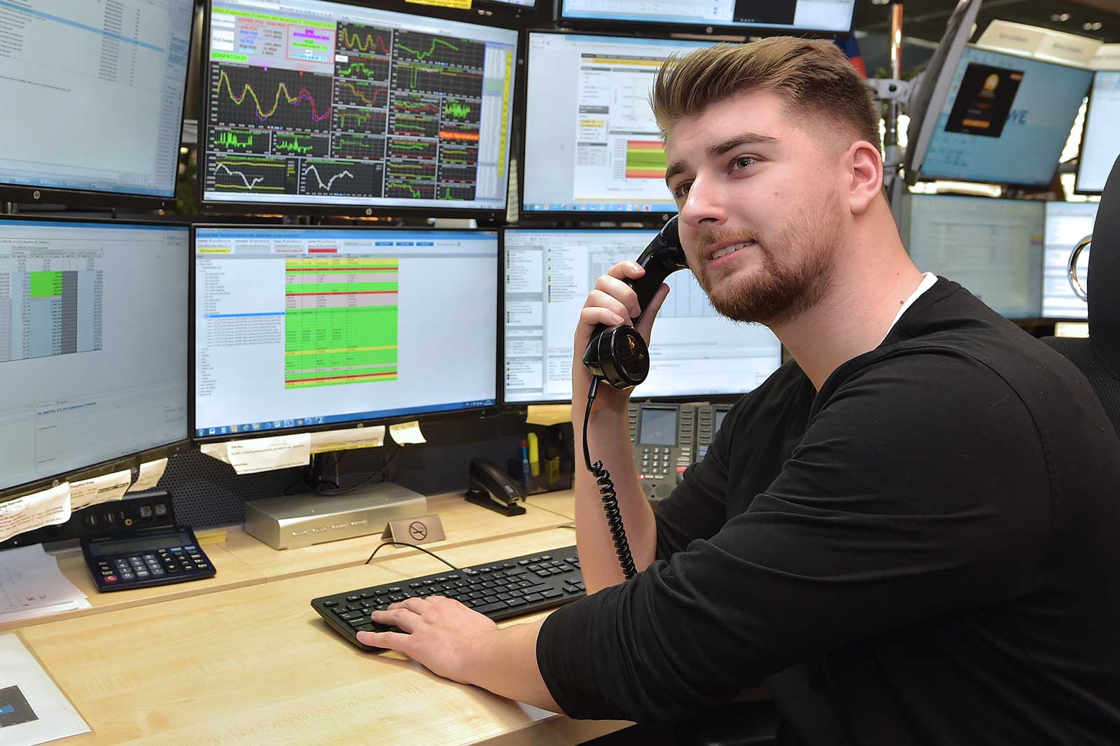 A trader using a phone in front of multiple computer screens displaying financial graphs and data tables.