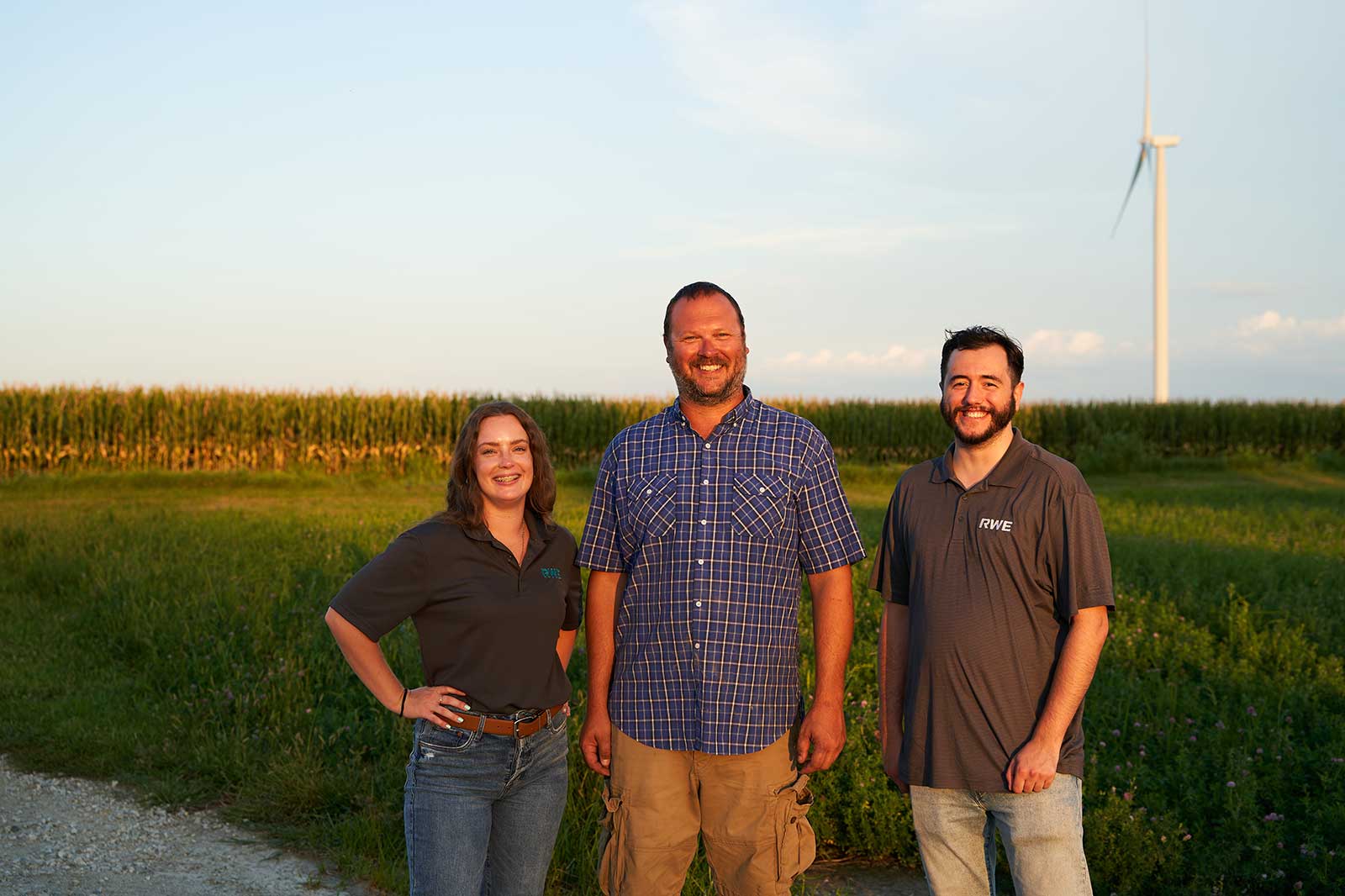 Three individuals stand in a field with corn crops and a wind turbine in the background, showcasing a rural landscape.