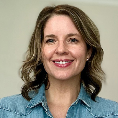 A woman with wavy, shoulder-length hair wearing a denim shirt against a neutral background.