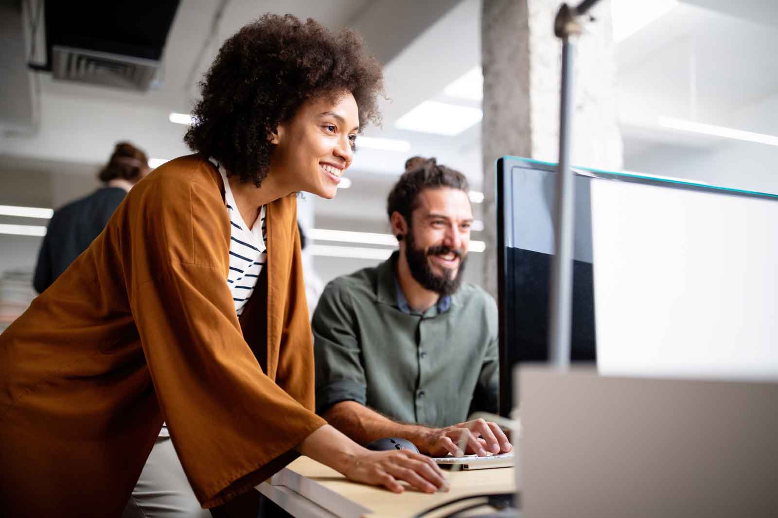 A person in a brown cardigan leans over a desk to assist another individual using a computer in a modern office environment.