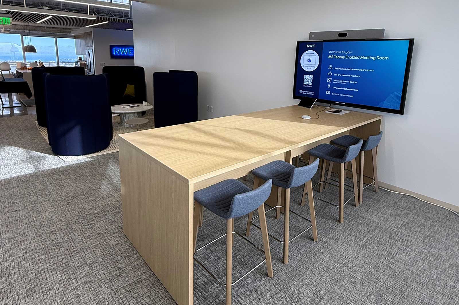A modern meeting room with a wooden table and blue chairs, featuring a large screen displaying MS Teams information.