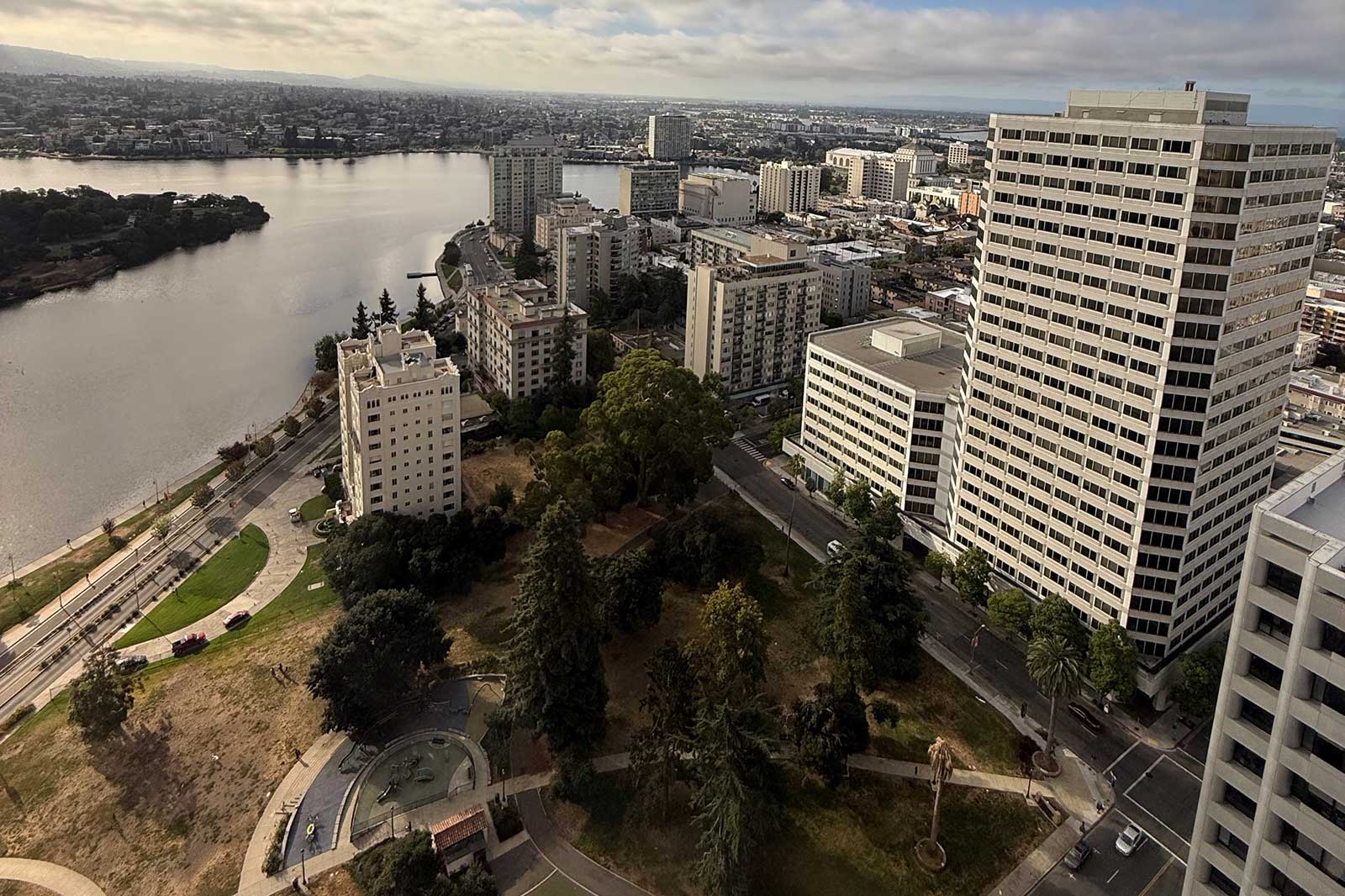 Aerial view of a cityscape with buildings beside a tranquil river and green parks, under a cloudy sky.