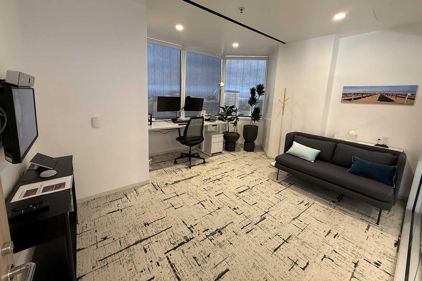 A modern office with two computer monitors, a grey sofa, and potted plants in a bright room with large windows.