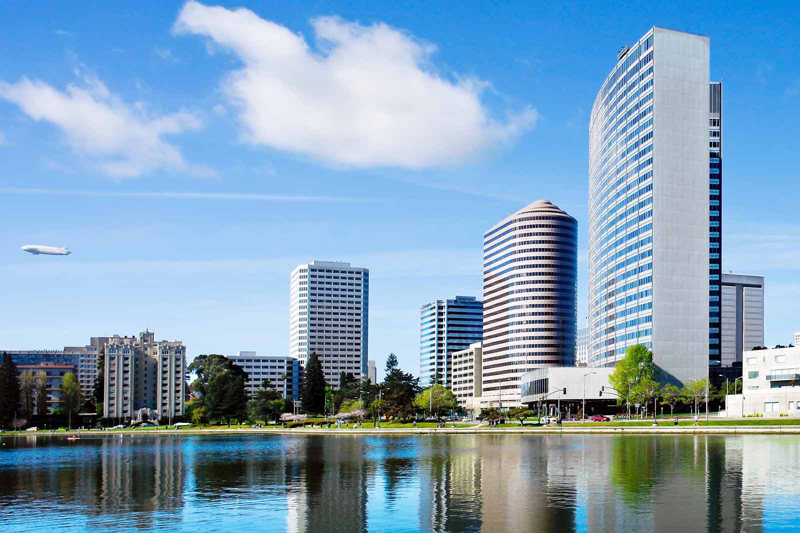 A scenic urban landscape featuring modern skyscrapers reflected in water, with fluffy clouds and a blimp in the sky.