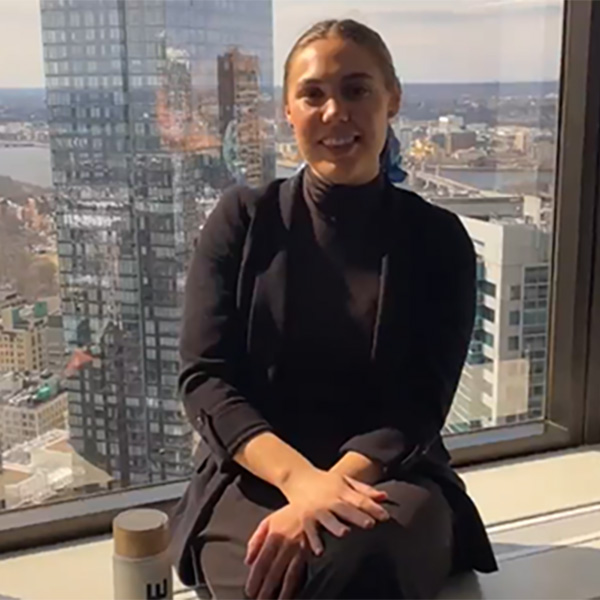 A woman in a black outfit sits by a large window overlooking a city skyline, with buildings and a river visible in the background.