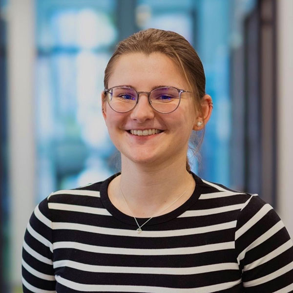A woman with hair tied back wears a black and white striped shirt. She stands in a professional setting.