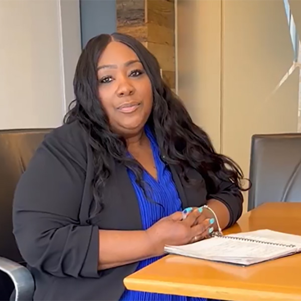 A woman with long black hair is seated at a table, wearing a black blazer over a blue top, with a notebook in front of her.