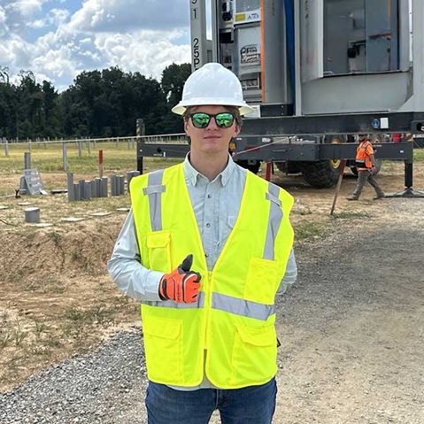 A construction worker in a bright yellow safety vest and white hard hat gives a thumbs up on a job site with machinery.