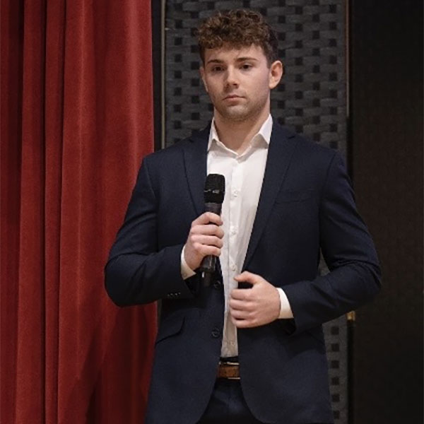 A man in a smart suit holding a microphone, standing in front of red curtains, appearing engaged in a speech.