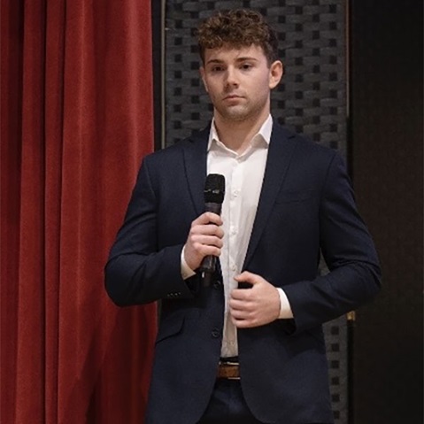 A man in a smart suit holding a microphone, standing in front of red curtains, appearing engaged in a speech.