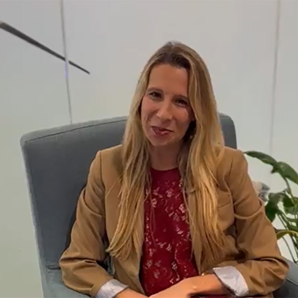 A woman with long hair sits in a gray armchair, wearing a brown blazer over a red blouse, with green plants in the background.