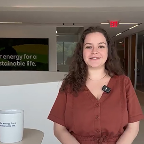 A person in a brown outfit stands in a modern office, with a coffee mug and sustainability message in the background.