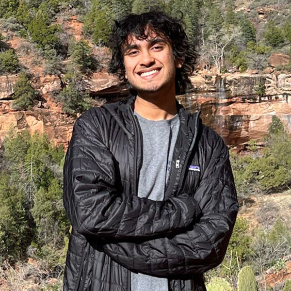 A person wearing a black jacket and grey t-shirt stands with arms crossed in front of a rocky, forested landscape.