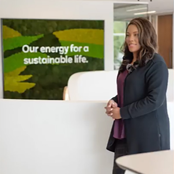 A woman stands in an office space beside a green wall with the slogan 'Our energy for a sustainable life' displayed prominently.