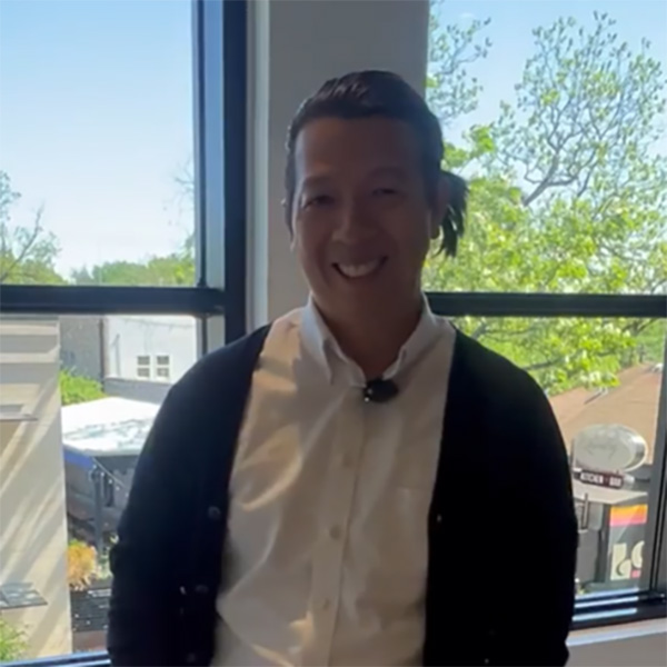 A person wears a black cardigan over a white shirt, standing by a large window with greenery and buildings outside.