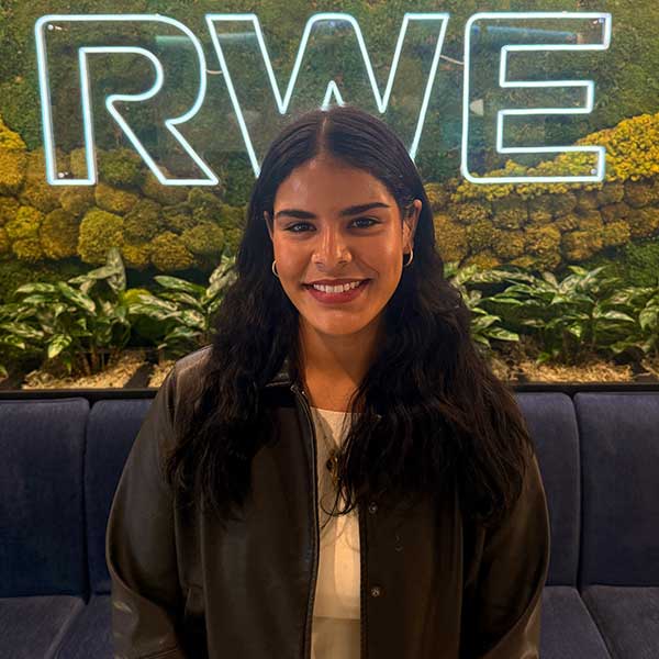 A person stands in front of a green wall with a glowing 'RWE' sign and a seating area featuring plants.
