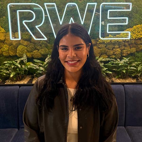 A person stands in front of a green wall with a glowing 'RWE' sign and a seating area featuring plants.